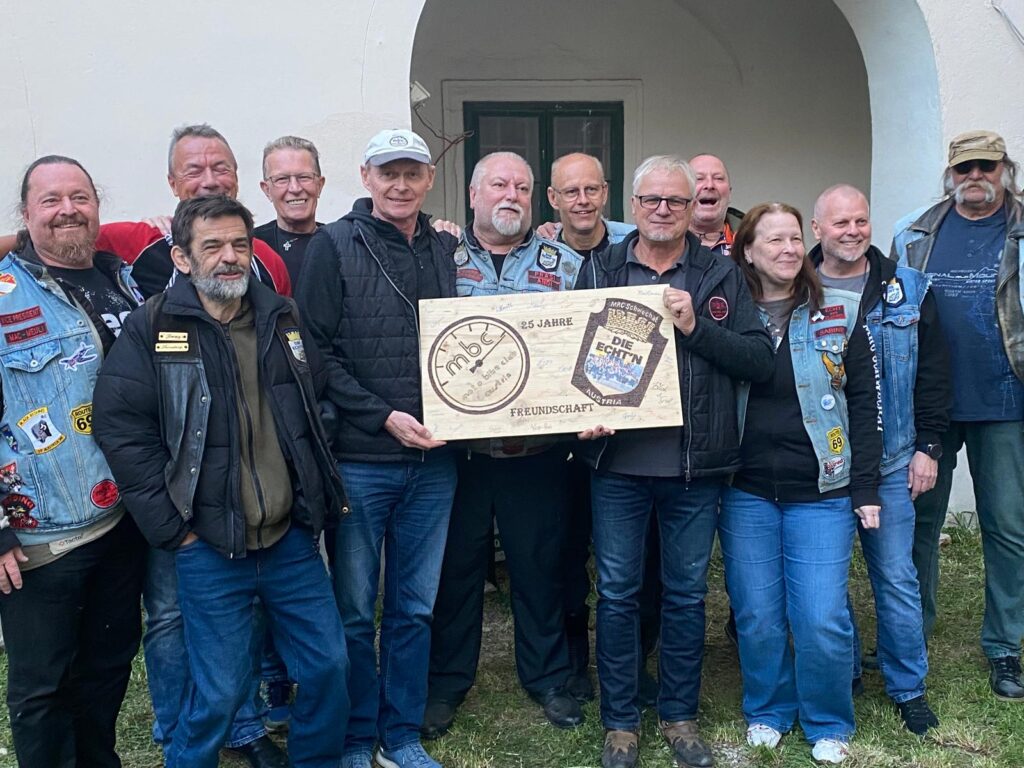 Gruppe von Mitgliedern des Moto Bike Club Austria (MBC) + Motorradclub "die Echt'n" posiert zusammen für ein Gruppenfoto, während sie ein Schild zum 25-jährigen Freundschaft präsentieren.