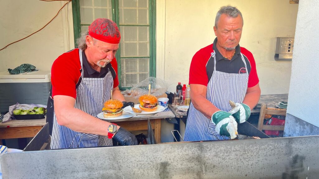 Zwei Männer bereiten Burger und Grillgerichte in einer Küche im Freien vor. Einer trägt eine rote Rabattmütze und hält einen Teller mit Burger, der andere verwendet Handschuhe und wendet das Grillgut.